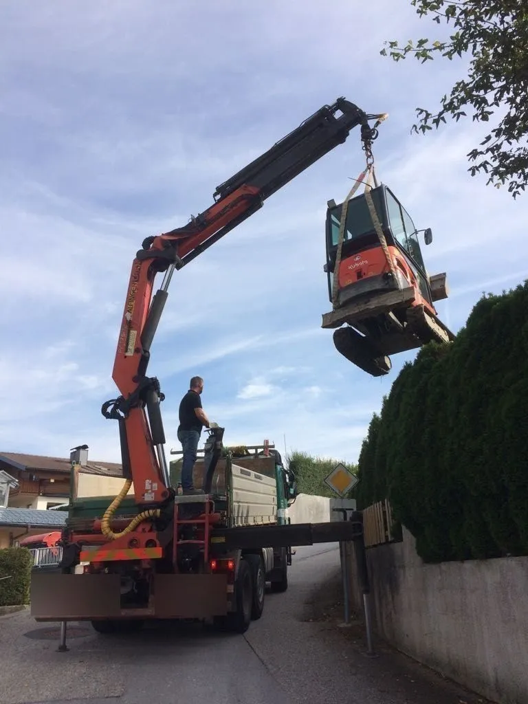Ein Ladekran hebt einen Minibagger über eine hohe Hecke